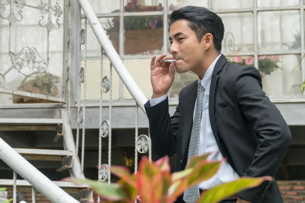 A young, Asian man in a suit smoking outside, next to a spiral staircase