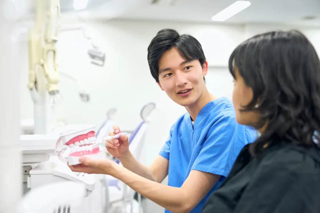 A young, male dentist in blue scrubs showing a patient a model of the mouth