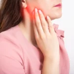 Woman in pink shirt pressing her hand against her aching jaw, a sign that she needs wisdom tooth removal in Singapore